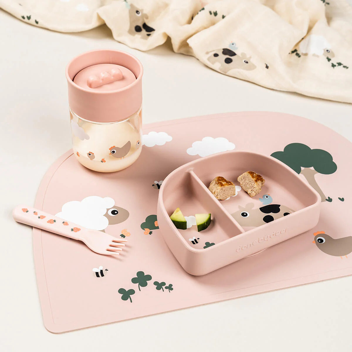 A Done by Deer set including 360 learning cup with milk in it, table mat, compartment plate containing snacks and fork all in powder pink and set on a white surface with Done by Deer printed fabric in the background.