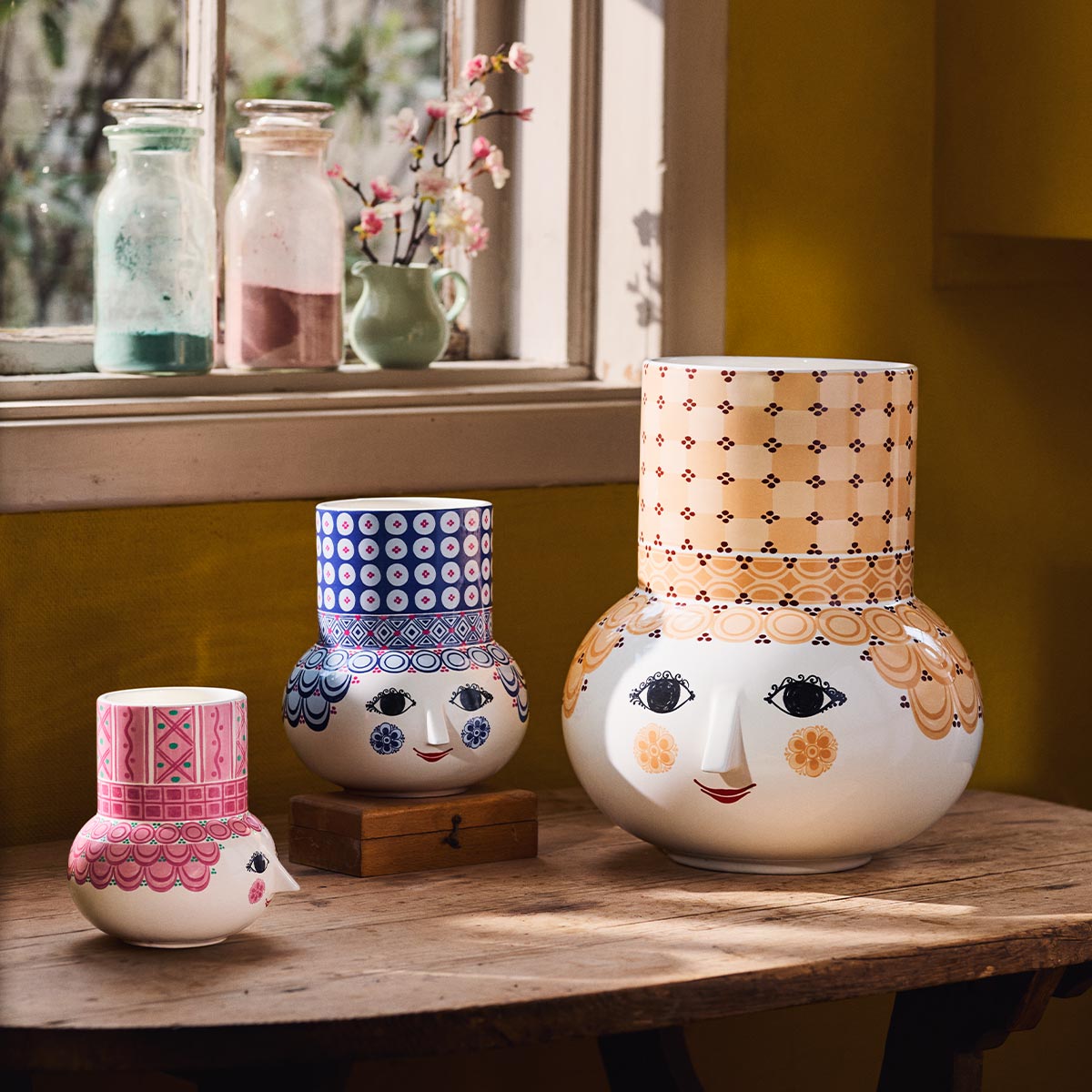 Three decorative Bjørn Wiinblad Olga vases with face designs on a wooden surface, with a window and plants in the background.
