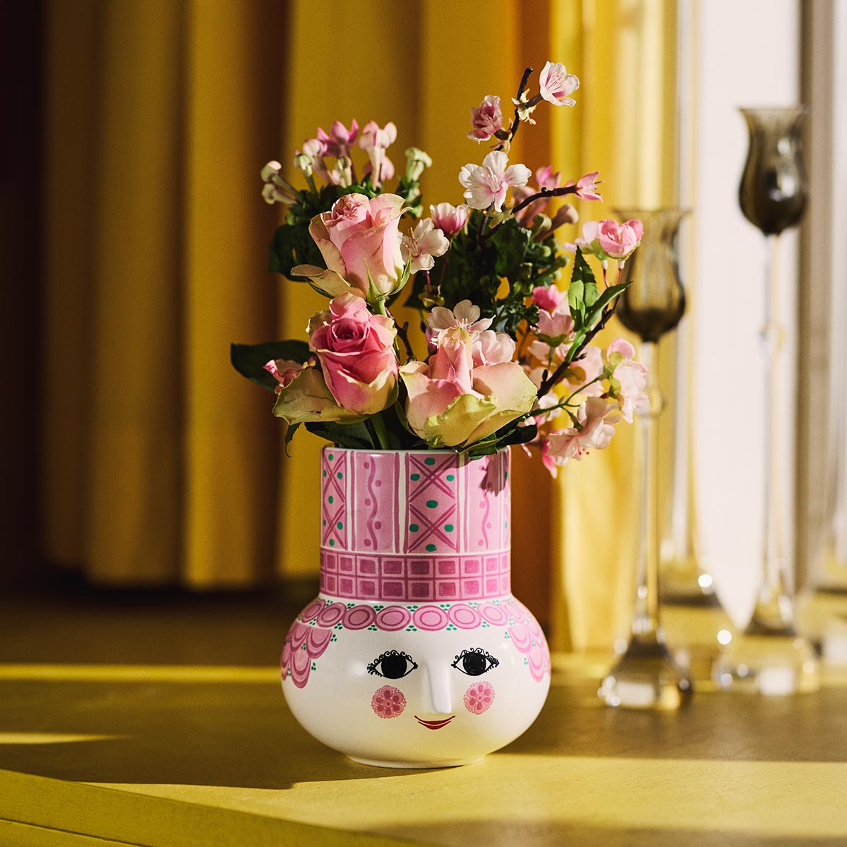 Decorative Bjørn Wiinblad Olga pink vase with face design and pink floral arrangement on a wooden surface.