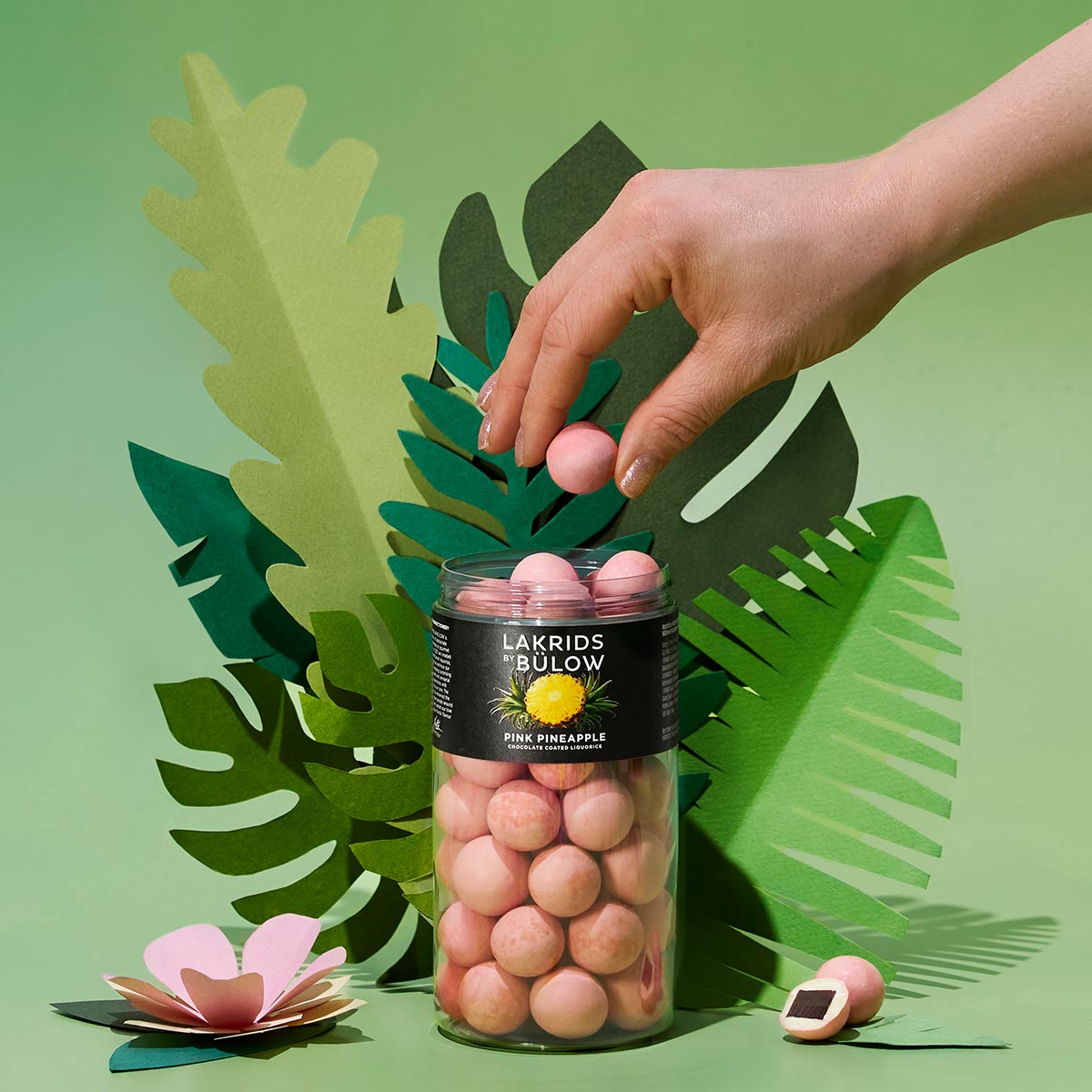 Hand reaching for pink pineapple LAKRIDS BY BÜLOW in a glass jar with tropical leaves on a green background