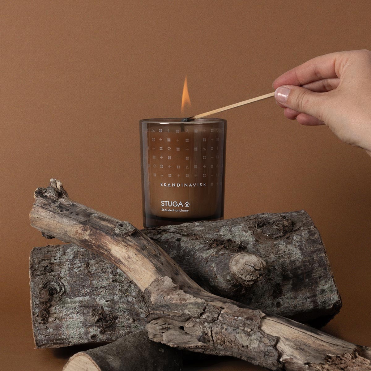 Hand lighting a Skandinavisk candle labeled Stuga with a wooden match against a brown background.