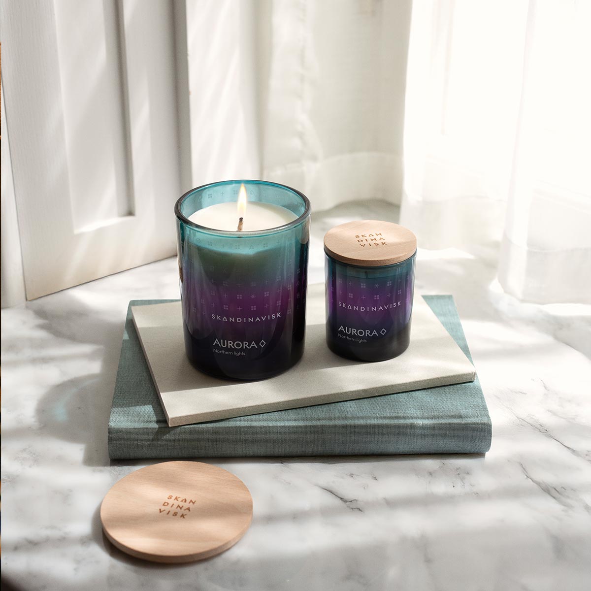 Two gradient Skandinavisk candles labeled 'Aurora' on a marble surface with a white curtain background.