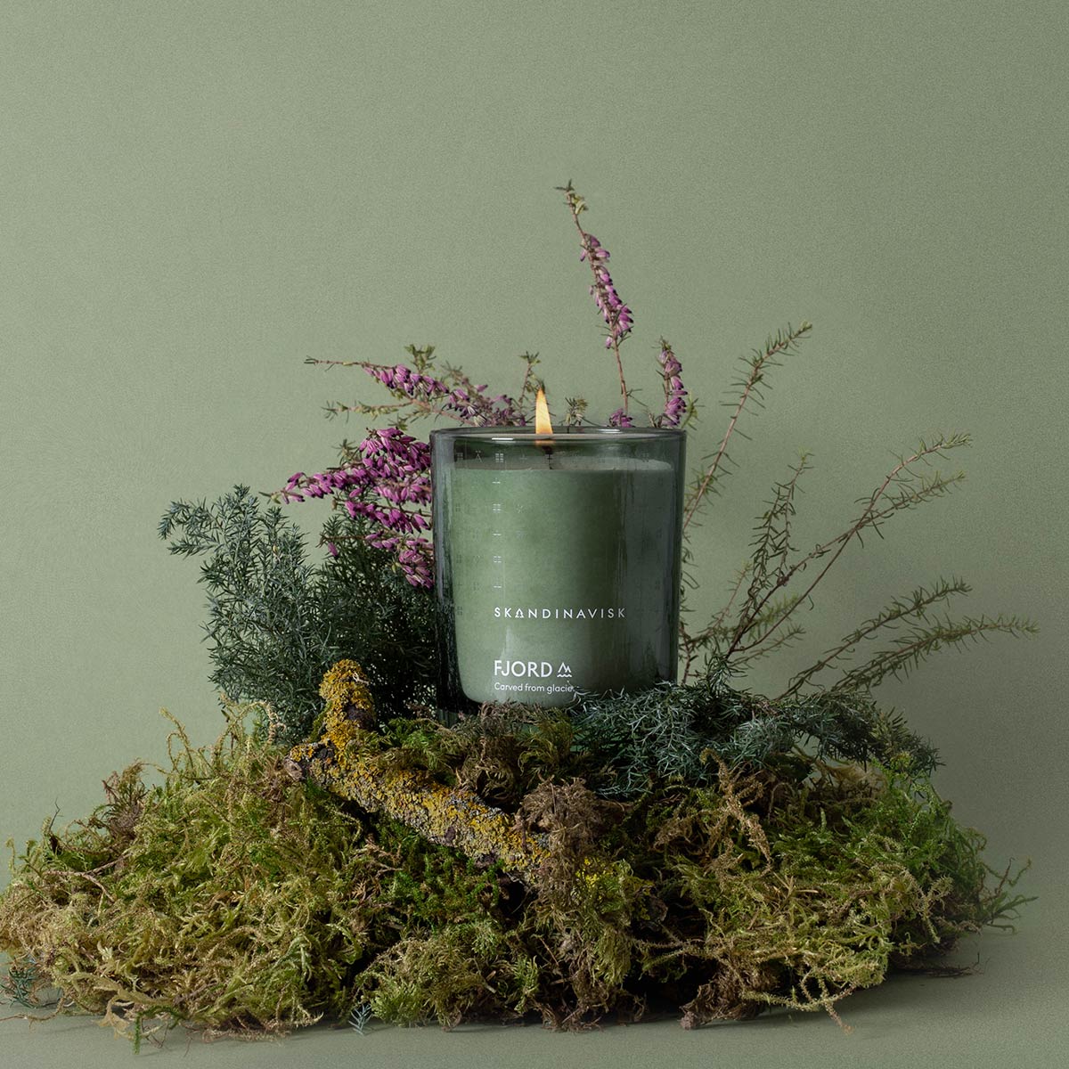 Candle with 'Skandinavisk' branding on a bed of moss and plants against a green background