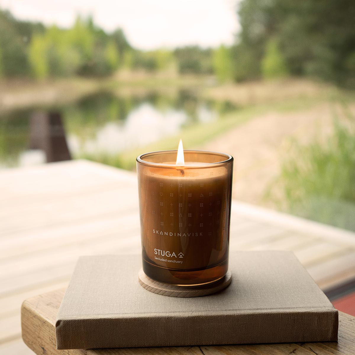 Candle in a glass container labeled 'STUGA' on a wooden surface with a blurred natural background