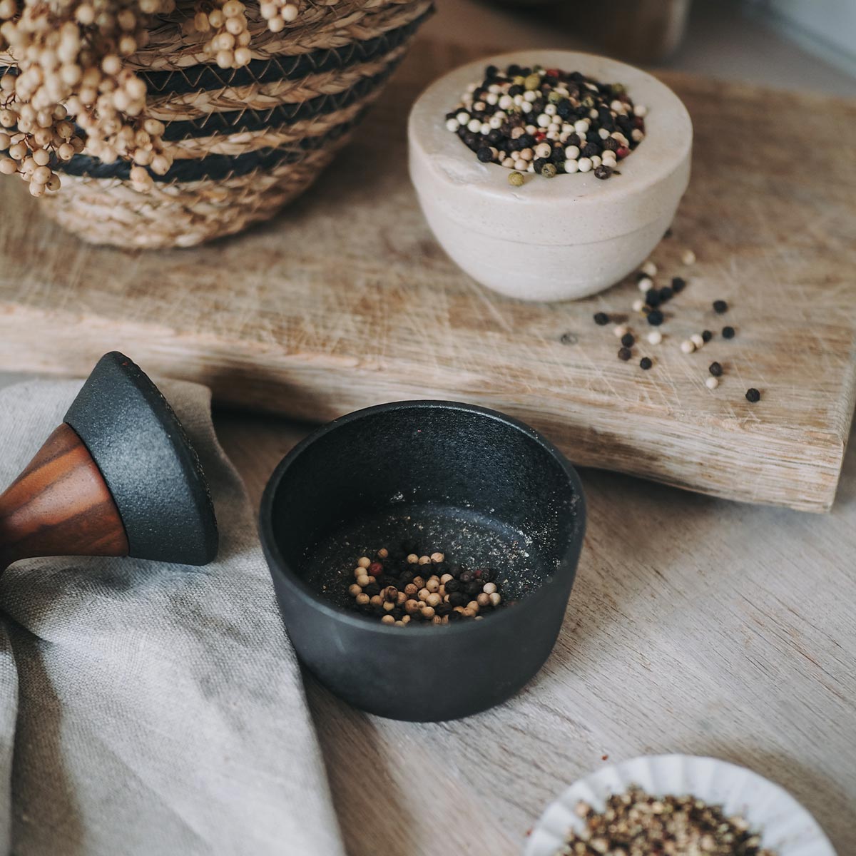 Black Crushgrind metal mortar with peppercorns on a wooden surface with a pestle and a cloth and woven basket and additional bowls of peppercorns.