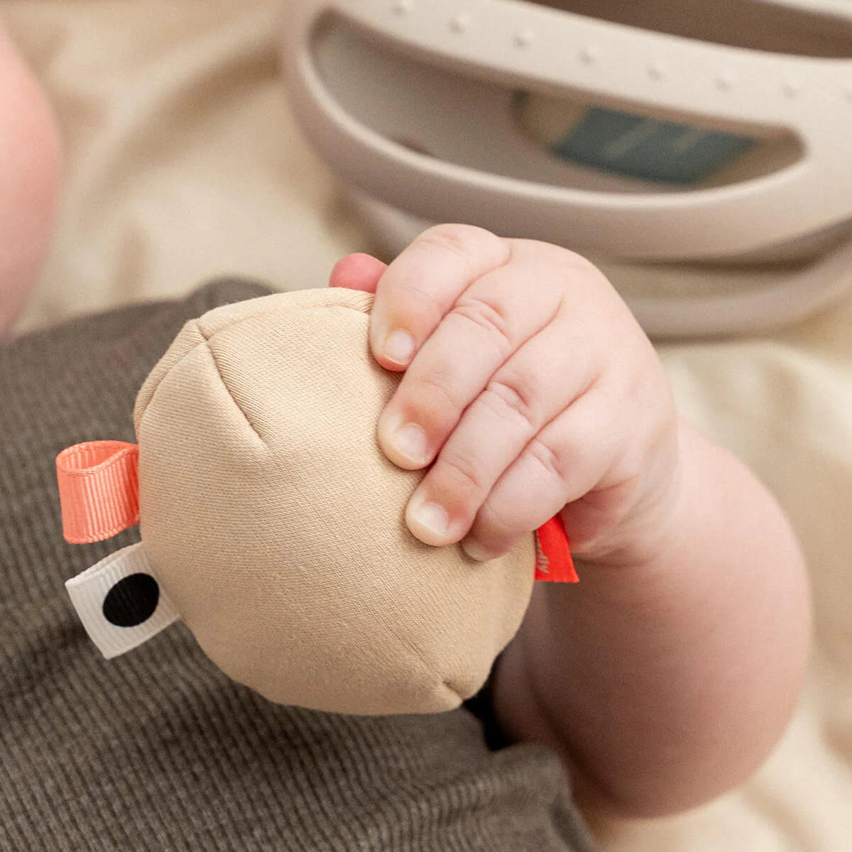 Child's hand holding the egg rattle from Done by Deer 'Clucky' teether rattle with the teether part of the toy shown in the background.