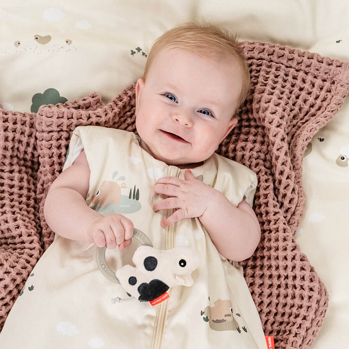 Smiling baby lying on a pink blanket with a 'Dotti' Done by Deer sensory rattle teether.