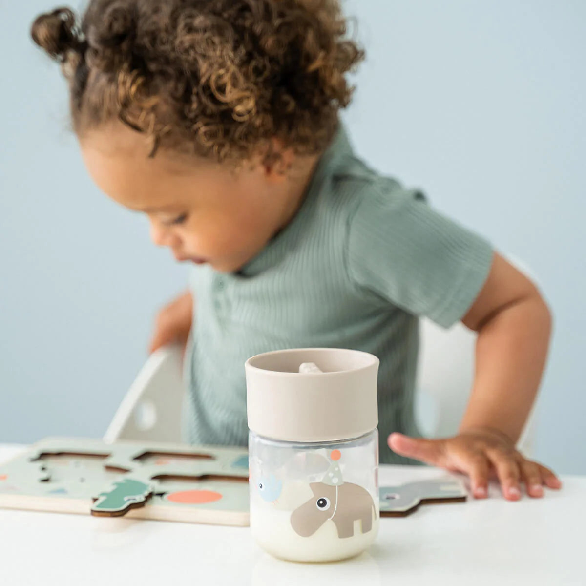 A child sitting at a white table with a Done by Deer 360 drinking cup in the colour sand next to a wooden puzzle.