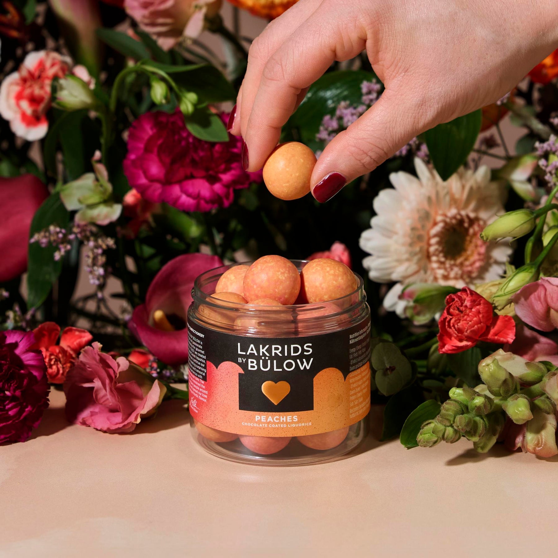 Hand holding a peach over a jar of Lakrids by Bülow peaches with flowers in the background