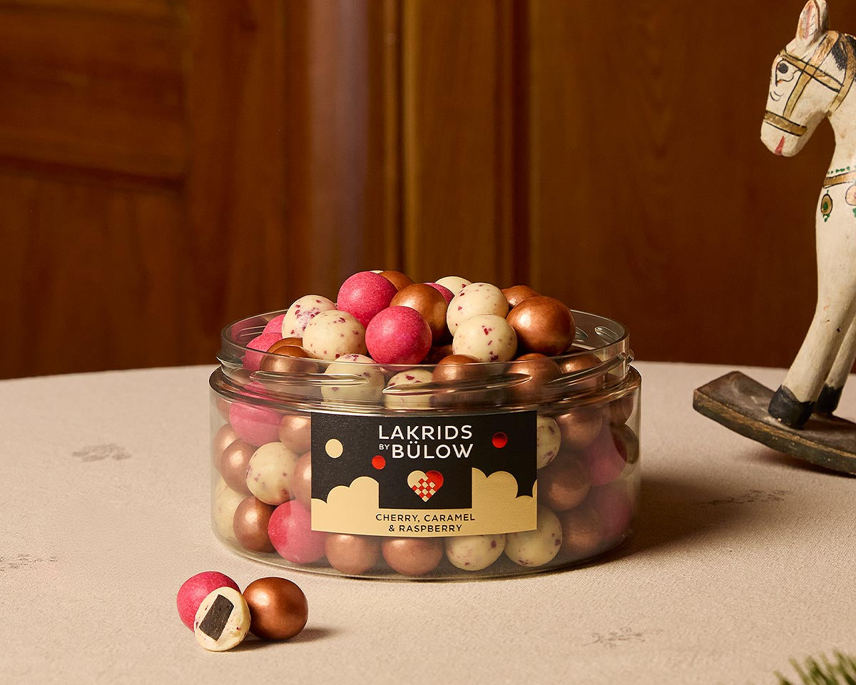 An open clear plastic jar filled with assorted gold, red and white speckled 'LAKRIDS BY BÚLOW' in cherry, caramel and raspberry flavours against on a table with a cream tablecloth next to a sprig of fir and a rocking horse ornaments
