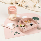 A Done by Deer set including 360 learning cup with milk in it, table mat, compartment plate containing snacks and fork all in powder pink and set on a white surface with Done by Deer printed fabric in the background.