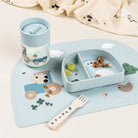 Done by Deer 360 drinking cup, table mat, fork and plate, all in blue, on a white table with Done by Deer printed fabric in the background.