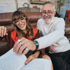 Two people wearing Izipizi glasses sitting together, one holding a book, in a cozy indoor setting.