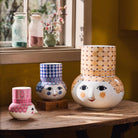 Three decorative Bjørn Wiinblad Olga vases with face designs on a wooden surface, with a window and plants in the background.