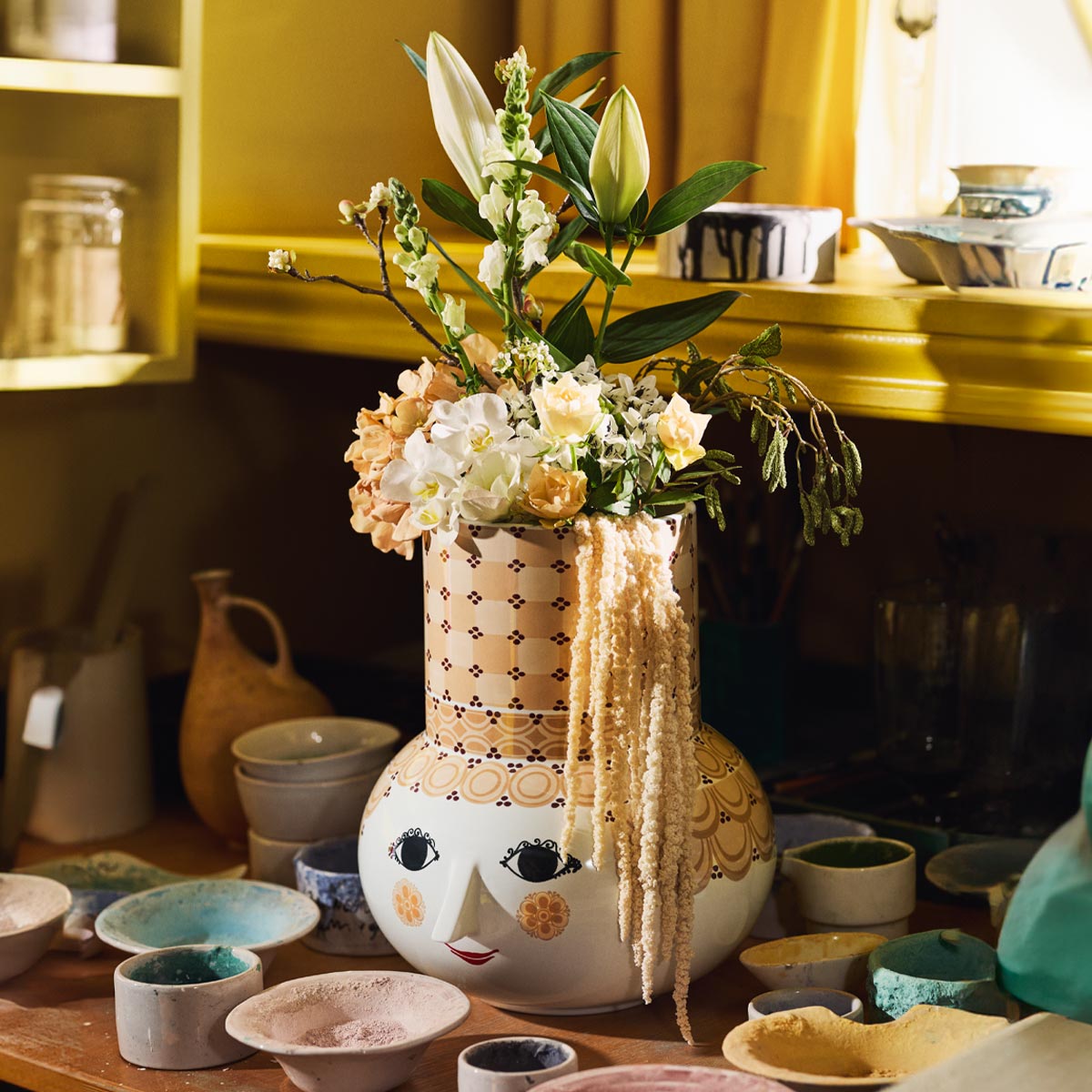 Decorative Bjørn Wiinblad  Olga sand vase with face design and floral arrangement on a table with various ceramic items.