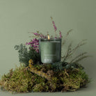 Candle with 'Skandinavisk' branding on a bed of moss and plants against a green background