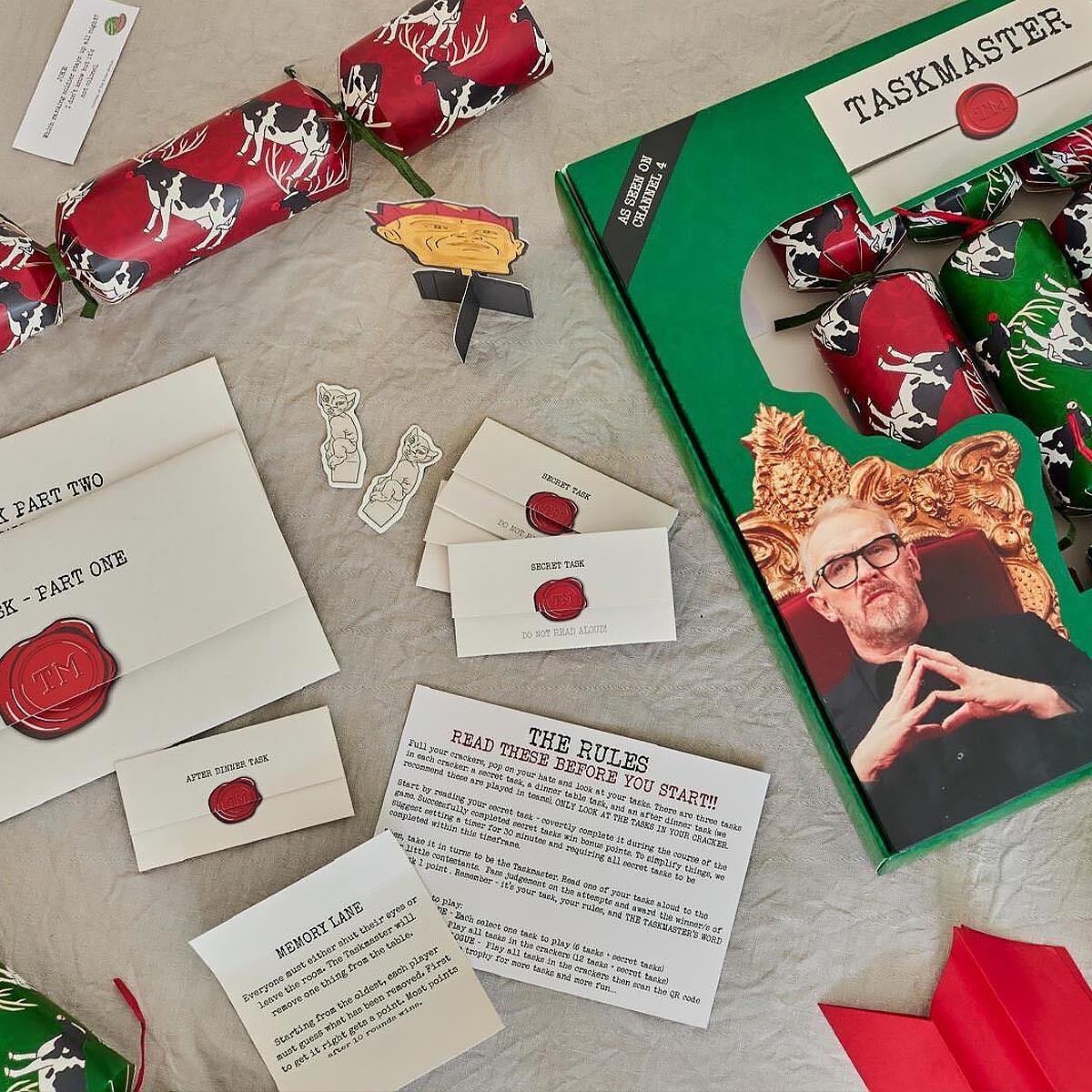 Taskmaster Game components including cards, envelopes, and a box with a person's face on it, on a textured surface.