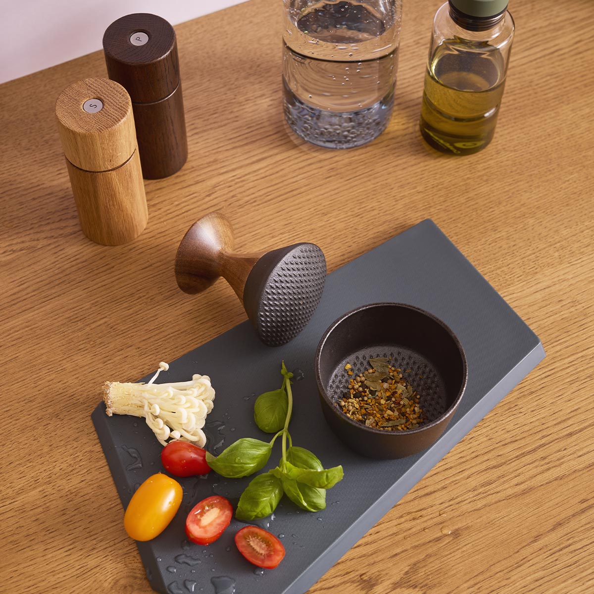 Crushgrind pestle and mortar on chopping board with vegetables herbs and seeds on a wooden surface with salt and pepper grinders and oil