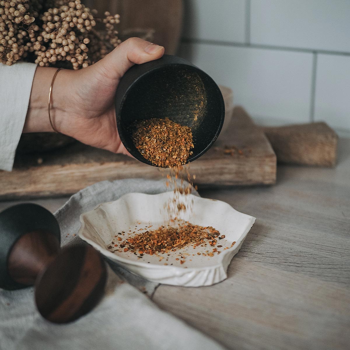 Person pouring ground herbs into a bowl from a Crushgrind metal mortar with a pestle and a cloth on a wooden surface.