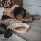 Person pouring ground herbs into a bowl from a Crushgrind metal mortar with a pestle and a cloth on a wooden surface.