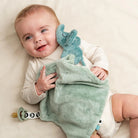 Baby holding a Done by Deer 'Elphee' comfort blanket with pacifier attachment lying on a beige blanket.