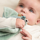 Close up of a bay holding a Done by Deer 'Elphee' comfort blanket with pacifier attachment to their mouth.