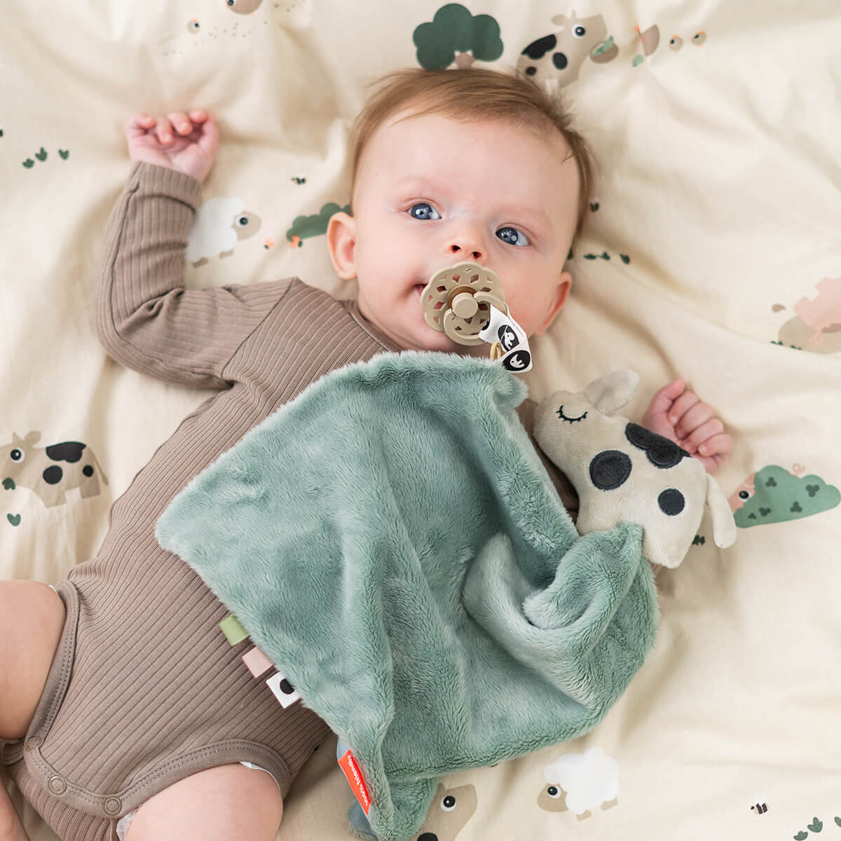 Baby with pacifier in their mouth which is attached to a Done by Deer 'Dotti' comfort blanket whilst lying on matching bedding.