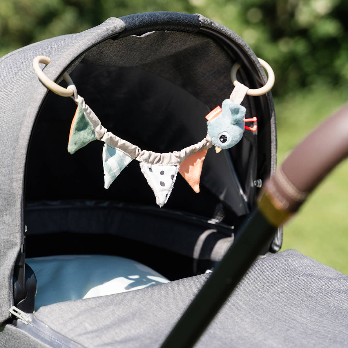 A grey pram in a grassy setting with Done by Deer 'Celebration' pram toy hanging from the hood.