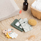 A selection of Done by Deer branded products including the sensory activity ring in Happy Cloud, muslins, brush, sponge and toiletries scattered over a beige rattan rug. 