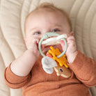 Baby holding a Done by Deer sensory activity toy in  the Happy Cloud design up to their mouth whilst lying on a beige blanket.