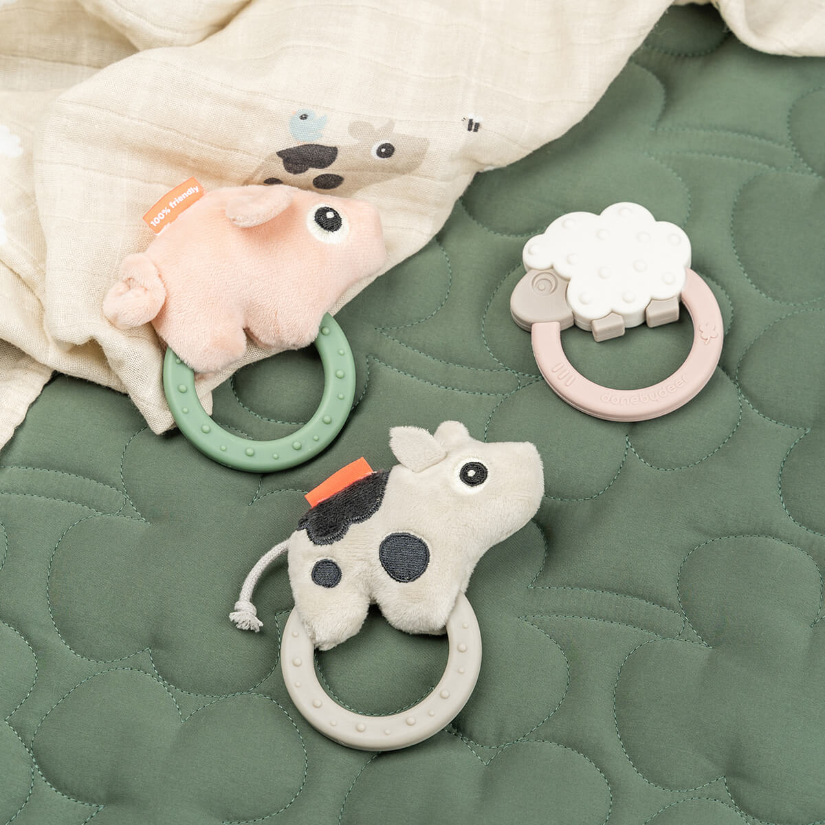 Three Done by Deer sensory rattle teethers and matching muslin blanket on a green quilted bedspread.