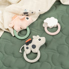 Three Done by Deer sensory rattle teethers and matching muslin blanket on a green quilted bedspread.