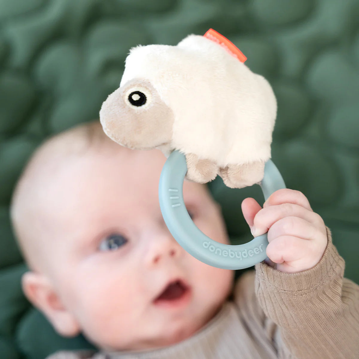 image from above of baby holding a 'Sheepy' sensory rattle and teether whilst lying on a green quilt.