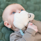 Baby holding a 'Sheepy' sensory rattle and teether by to their mouth whilst lying on a green quilt.
