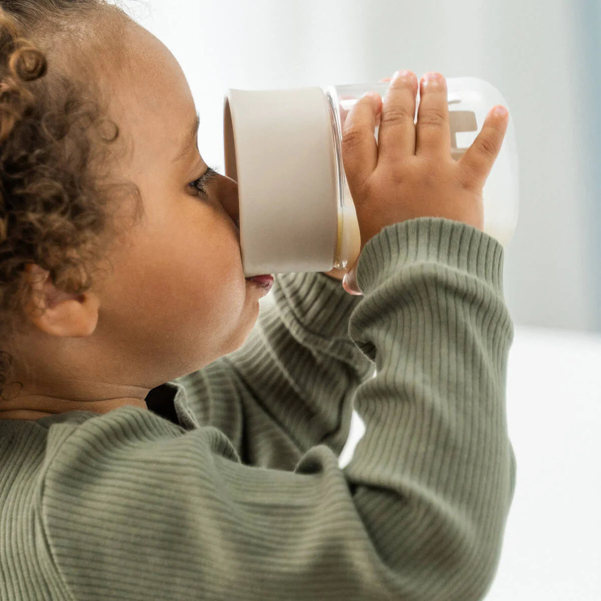 Photo taken from the side of a child with brown curls, wearing a green top and drinking from a Done by Deer drinking cup against a white backdrop.