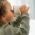 Photo taken from the side of a child with brown curls, wearing a green top and drinking from a Done by Deer drinking cup against a white backdrop.