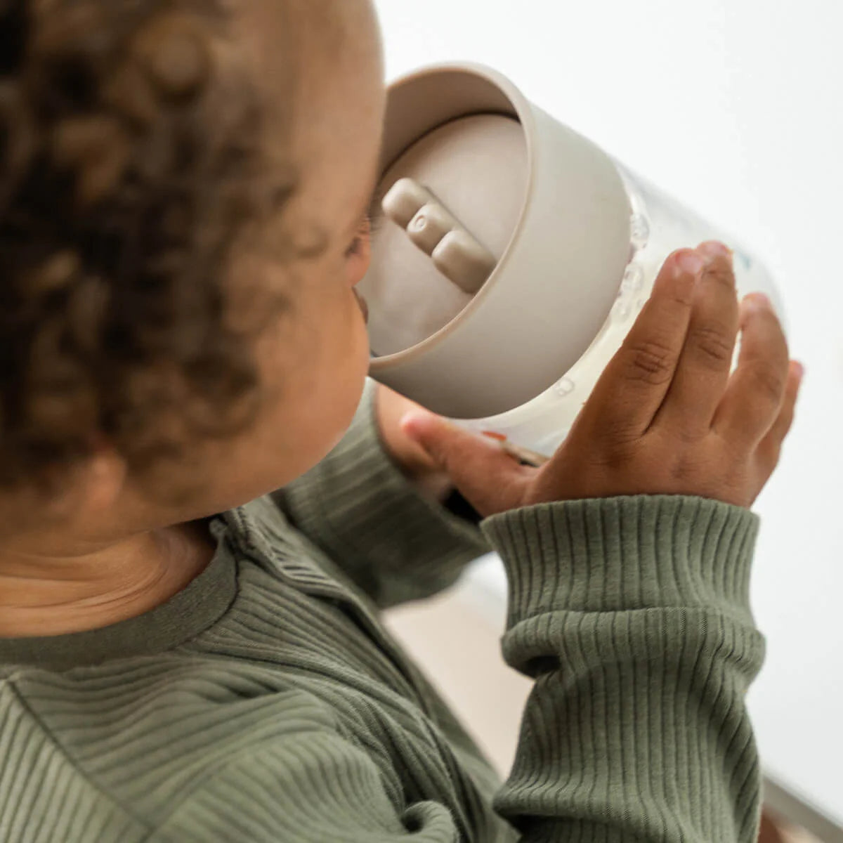 A child with brown curls, wearing a green top and drinking from a Done by Deer drinking cup against a white backdrop.