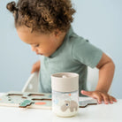 A child sitting at a white table with a Done by Deer 360 drinking cup in the colour sand next to a wooden puzzle.
