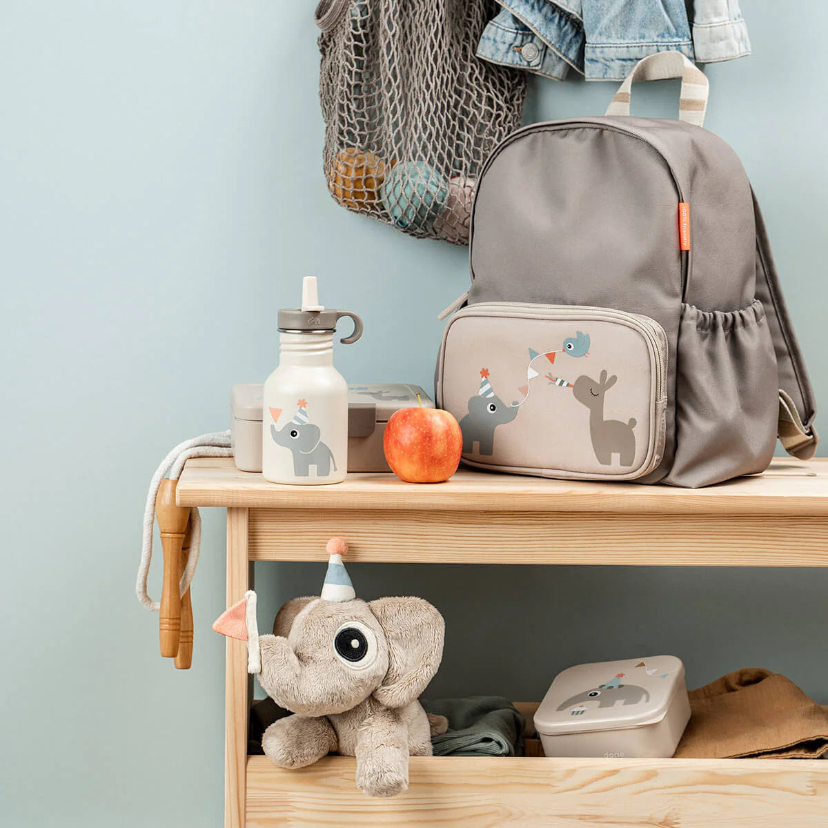 A wooden bench against a blue background featuring Done by Deer products in the colour 'sand' including a backpack, lunchbox, metal drinking bottle, sandwich box and soft toy. An apple sits on the bench and clothing and bags hang above.