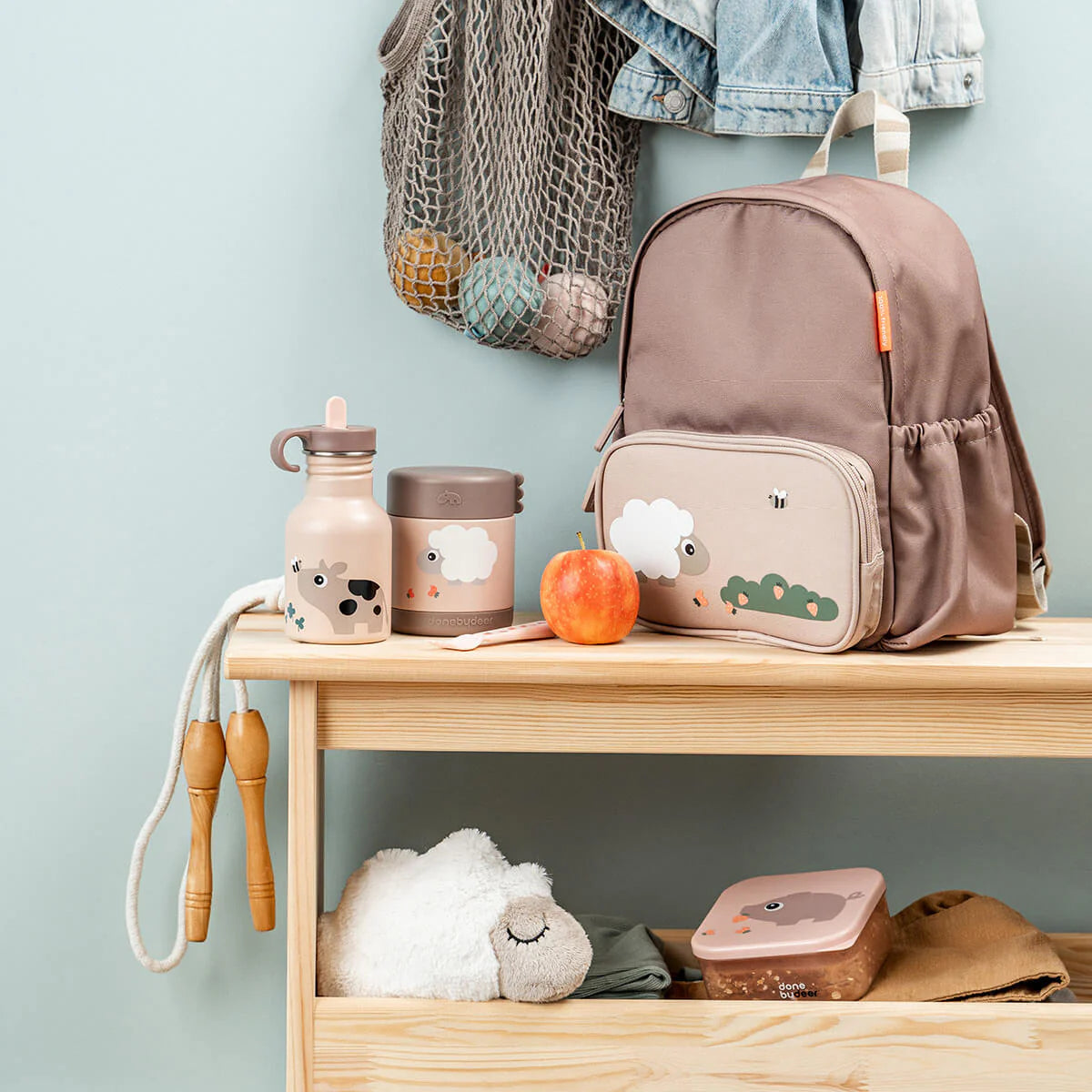 A wooden bench against a blue background featuring powder pink Done by Deer products including a backpack, lunchbox, metal drinking bottle, sandwich box and soft toy. An apple sits on the bench and clothing and bags hang above.