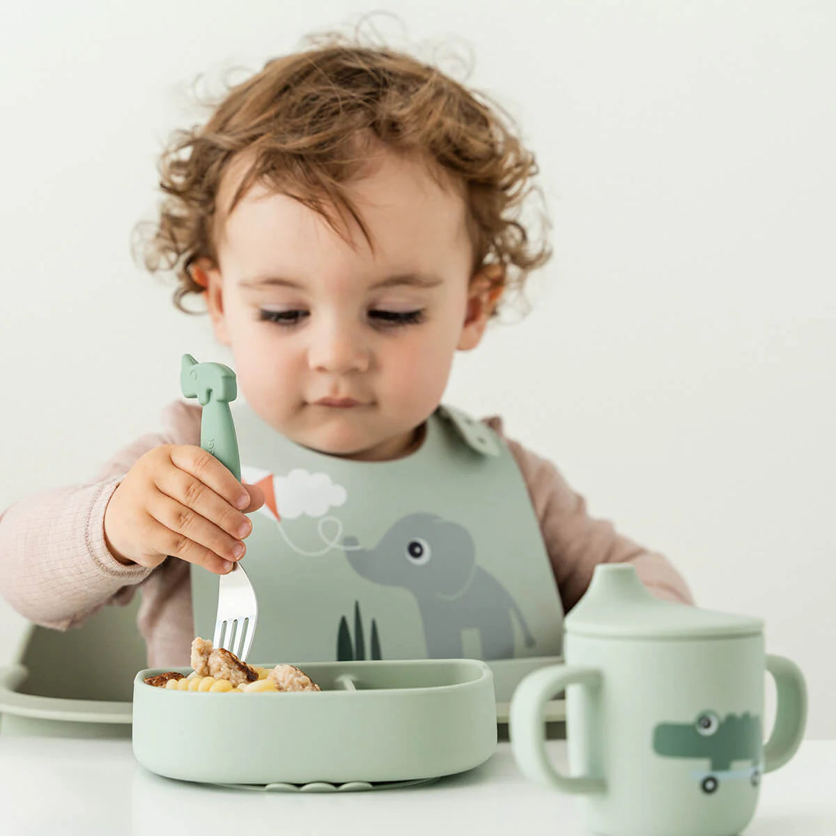 Child wearing a Done by Deer bib and eating from matching green tableware against a white background.