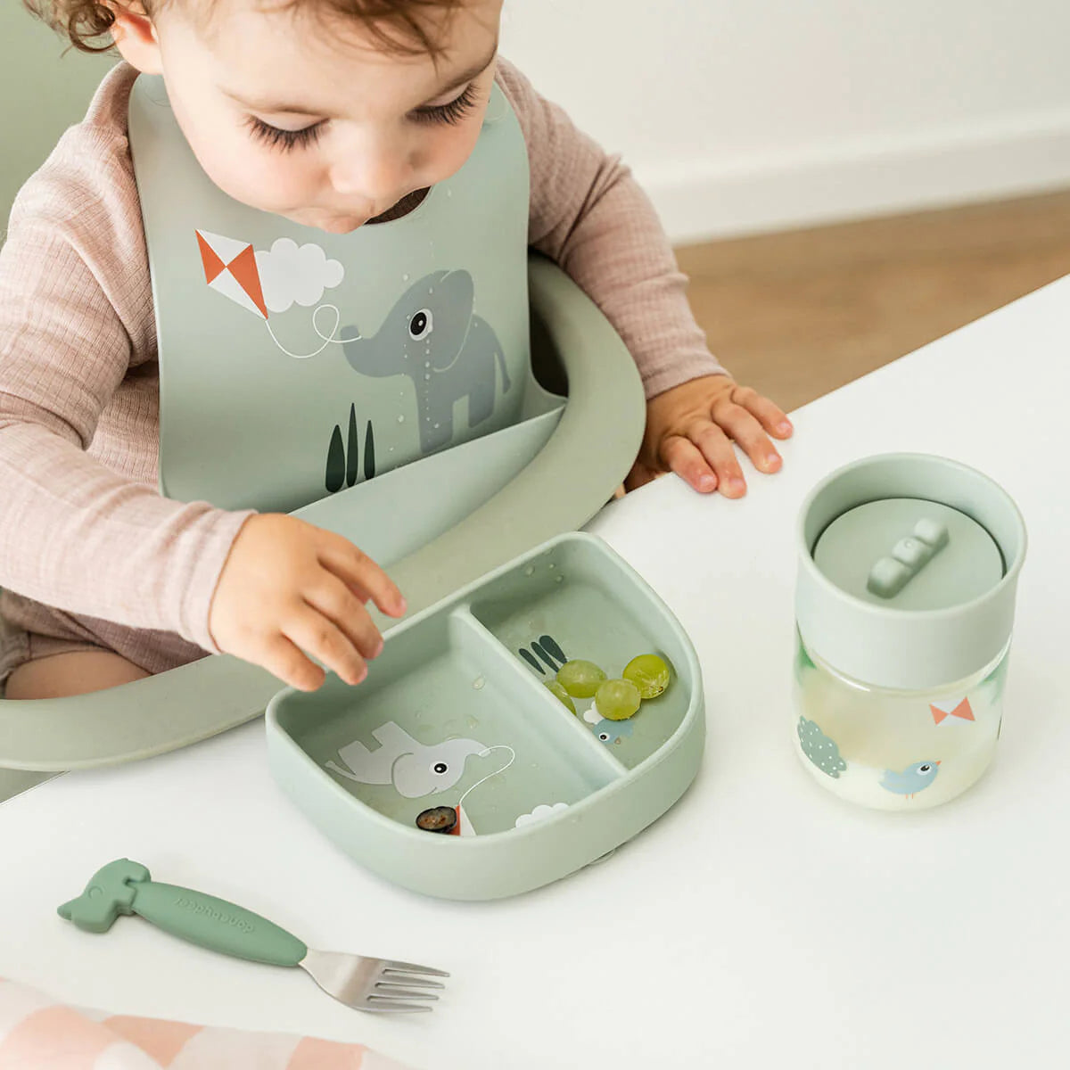 Toddler at a table wearing a Done by Deer bib and eating grapes from a plate with compartments. On the white table there is a 360 drinking cup and fork too. 