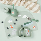 A photo from above with matching Done by Deer kids tableware including the silicone cup with straw, compartment plate, mat and cutlery with scattered blueberries on a pale sage coloured table. 