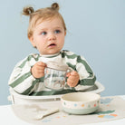 Child in a green and white striped bib holding a Done by Deer grippy spout cup behind a table featuring other pieces of tableware from the collection set against a light blue background.