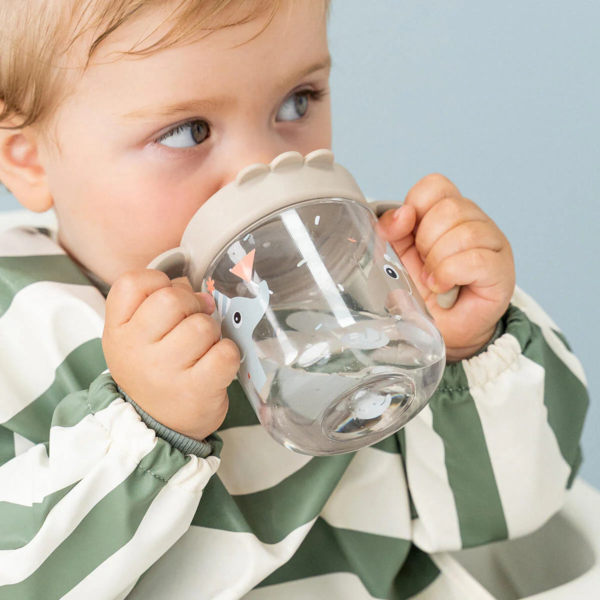 Child in striped green bib holding a Done by Deer grippy spout cup against a light blue background.