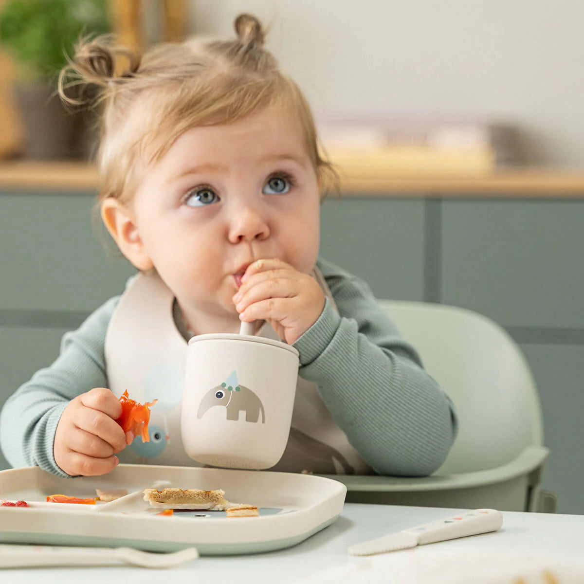 Child sat in a high chair drinking from a Done by Deer cup with silicone straw and eating from a matching compartment plate on a white table with cabinets and plants in the background.