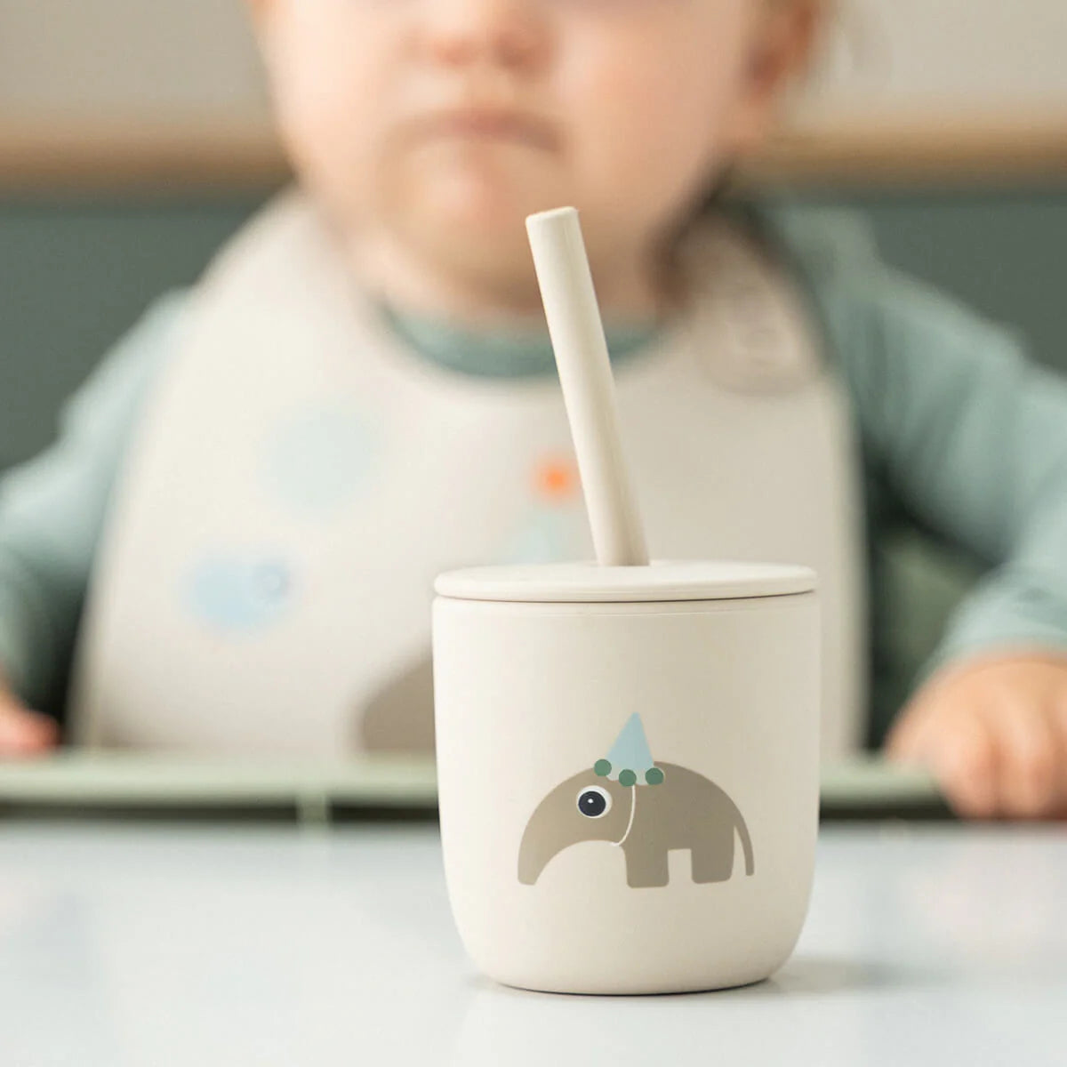 A Done by Deer silicone cup with straw on a white surface with a child sat in a high chair in the background.