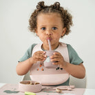 Child drinking from a pink Done by Deer cup with a straw, wearing a matching bib, sitting at a white table with a matching bowl, mat utensils and food scattered about against a white background.