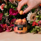 Hand holding a peach over a jar of Lakrids by Bülow peaches with flowers in the background