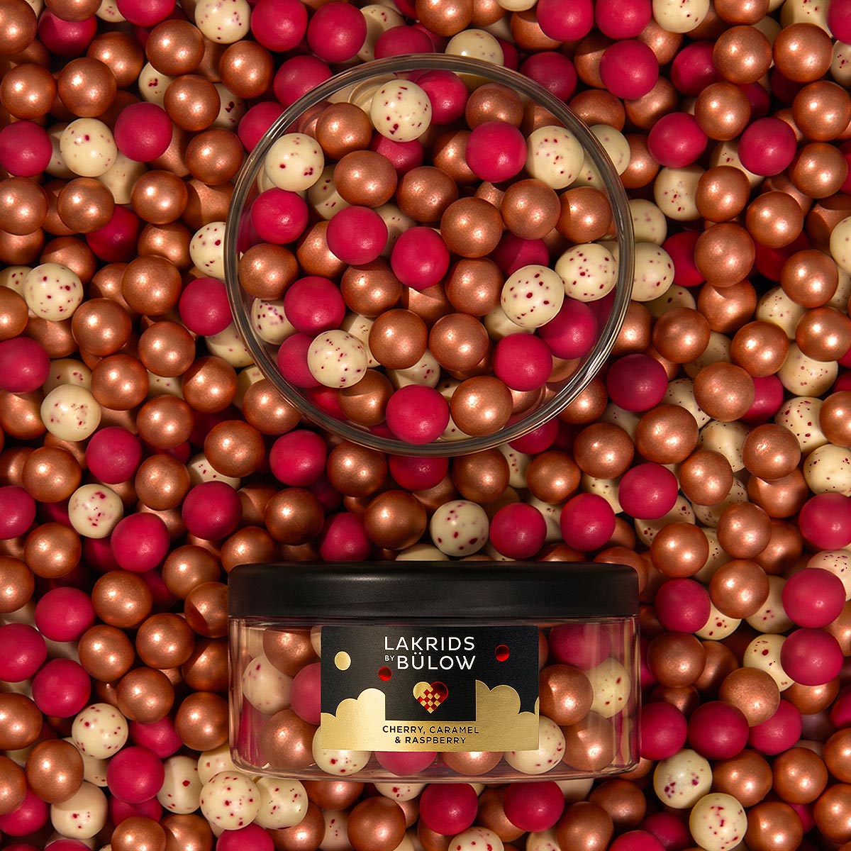 A clear plastic jar with a black lid filled with assorted gold, red and white speckled 'LAKRIDS BY BÚLOW' in cherry, caramel and raspberry flavours against a background filled with more of the gold, red and white speckled Lakrids confectionary.